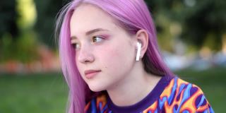 夏天的一天，一头粉红色头发、戴着耳机的潮女正在城市街道上使用智能手机。夏天的概念。Z一代的风格。社交媒体的概念。