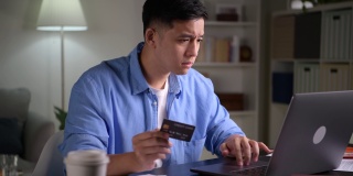 严肃的年轻亚洲男子，皱眉和担心，使用和看着信用卡，做网上购物，在家里用笔记本电脑进行网上支付交易。