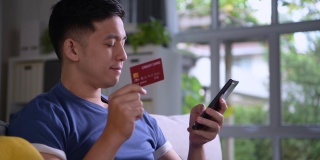 亚洲男子舒适地坐在家里，享受和浏览网上购物和方便快捷的支付使用信用卡在线支付和智能手机银行应用程序。