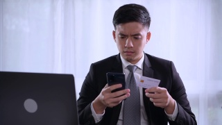 聪明的年轻亚洲商人坐在家里的办公室里，带着笔记本电脑，微笑着，用智能手机上的在线购物手机应用程序使用信用卡。