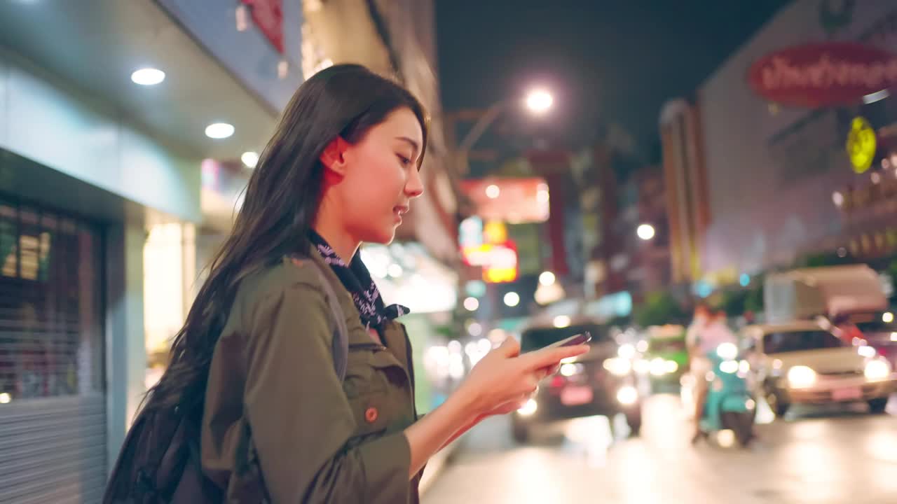 亚洲美丽的女人背包客滚动手机，而走在城市。有吸引力的女孩旅游旅行者独自在街上旅行，使用智能手机享受假期假期旅行的时间在夜间道路