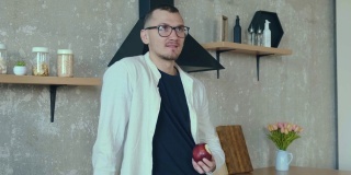 一个戴着眼镜的年轻人站在厨房边吃着苹果边用无线耳机和他的同事谈论工作项目。家的概念。移动电话。