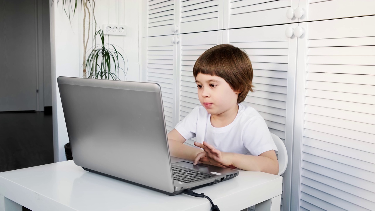 一个5岁的男孩坐在桌子旁，用笔记本电脑，看着屏幕。孩子在做作业，玩电子游戏，学习IT开发，看动画片。家庭远程在线教育理念