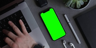 一个绿色屏幕的手机放在他的桌面上，正在笔记本电脑上打字