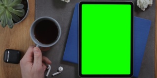 一名男子端着一杯咖啡，放在一台绿色屏幕的平板电脑旁边