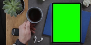 一名男子将一杯咖啡放在桌面上一个绿色屏幕的平板电脑旁边
