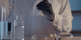 一个穿着晨衣、戴着防护橡胶手套的医生的特写镜头，从一个瓶子里收集液体，并把它倒进一个烧瓶里。科学实验室的研究工作。人类的手。