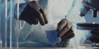 一名戴着防护橡胶手套的实验室助理在实验室设备的背景下搅拌一个烧瓶中的蓝色液体。制药行业。研究工作。
