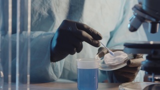 一位戴着防护橡胶手套的实验室助理在皮氏培养皿的棉垫上拍摄蓝色液体样本的特写镜头。在实验室设备的背景下。