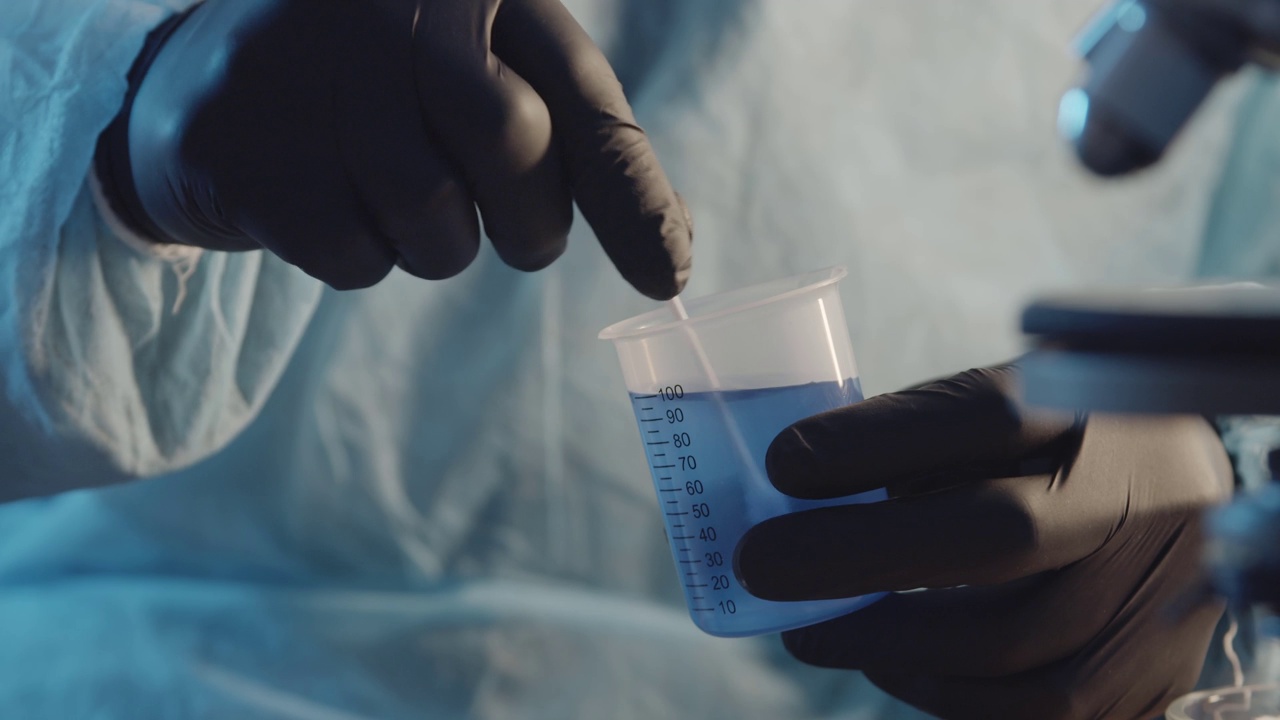 在实验室设备的背景下，一名戴着防护橡胶手套的实验室助理在一个测量杯中搅拌蓝色液体。制药行业。研究工作