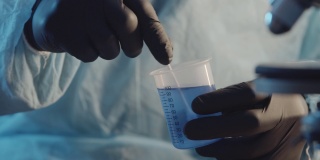 在实验室设备的背景下，一名戴着防护橡胶手套的实验室助理在一个测量杯中搅拌蓝色液体。制药行业。研究工作