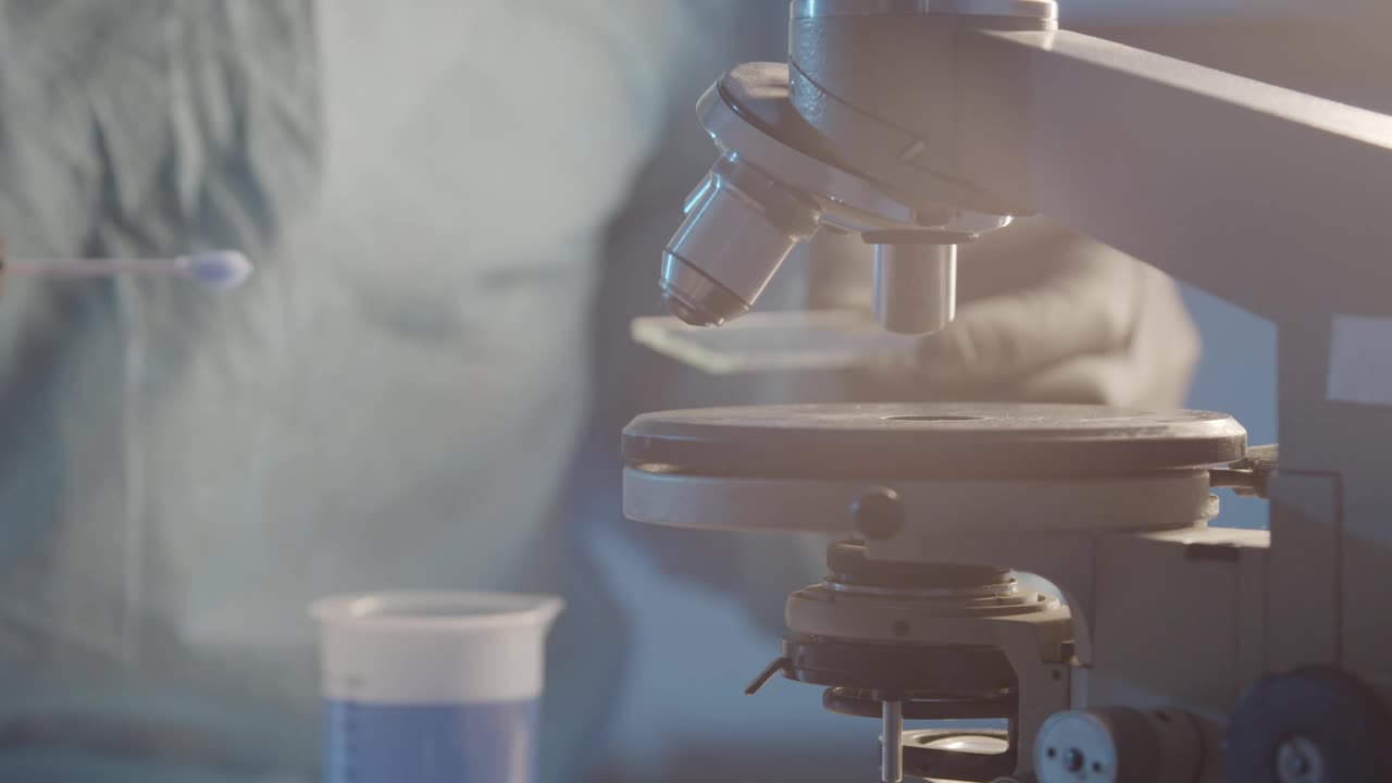 一名实验室的研究人员戴着防护橡胶手套，在实验室设备的背景下，将蓝色液体对准显微镜载玻片特写镜头。科学实验室的研究工作。