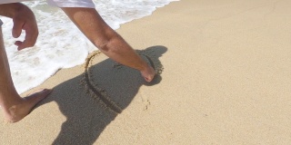 男人在沙滩上画着一颗心，海浪覆盖着一颗心的特写。浪漫的爱情象征在海边。爱，爱，心，画在沙滩上的沙