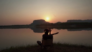 ฺ背影一个年轻人和休息在河边的山景日落。