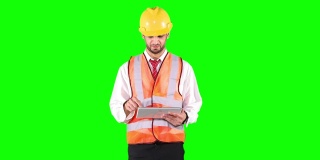 男性承包商戴着头盔，站在工作室的绿色屏幕背景下使用数字平板电脑