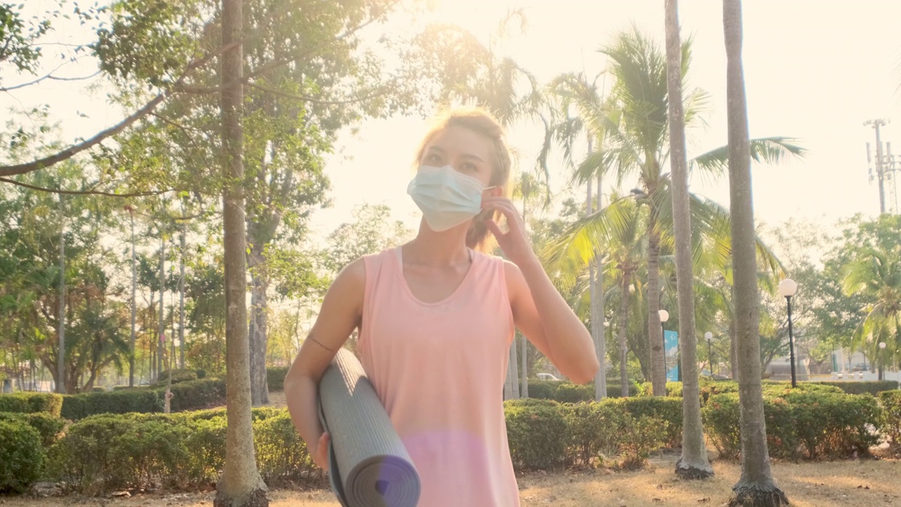 新常态健康生活理念。一名年轻的亚洲女性从COVID-19疫情中康复后，摘下医用口罩，在公园室外练习瑜伽。