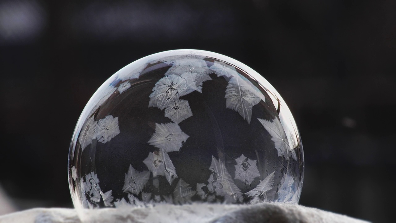 一个肥皂泡躺在黑色的雪地上，雪花落在上面结冰。冰气球模式