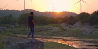 日落时分，在印度古吉拉特邦的万卡纳，一名散漫的印度男子走在河边，背景是山上的风车。选择性地聚焦于背景中的风车和山丘。