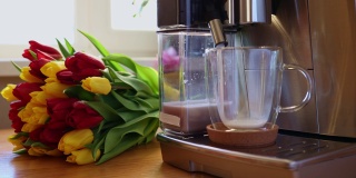 厨房的木桌上放着一杯咖啡、牛奶和郁金香花。早餐在咖啡机里现煮的卡布奇诺拿铁。时尚的家庭室内装饰。