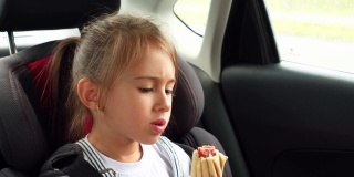 孩子在车里吃热狗。汽车后座上的女孩在旅途中被系上了安全带。