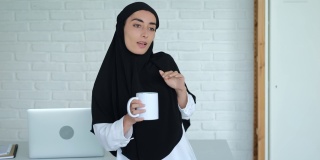 一名穆斯林妇女在辛苦工作后，在办公室的笔记本电脑前欢快地跳舞。一位戴着黑色头巾的美丽女子手里拿着一杯咖啡翩翩起舞。在办公室工作愉快吗