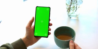 男人拿着带Chroma屏幕的智能手机，在咖啡馆的白色桌子上喝着卡布奇诺咖啡，通过互联网，滑动屏幕，使用网站，绿色屏幕的商家选择性聚焦