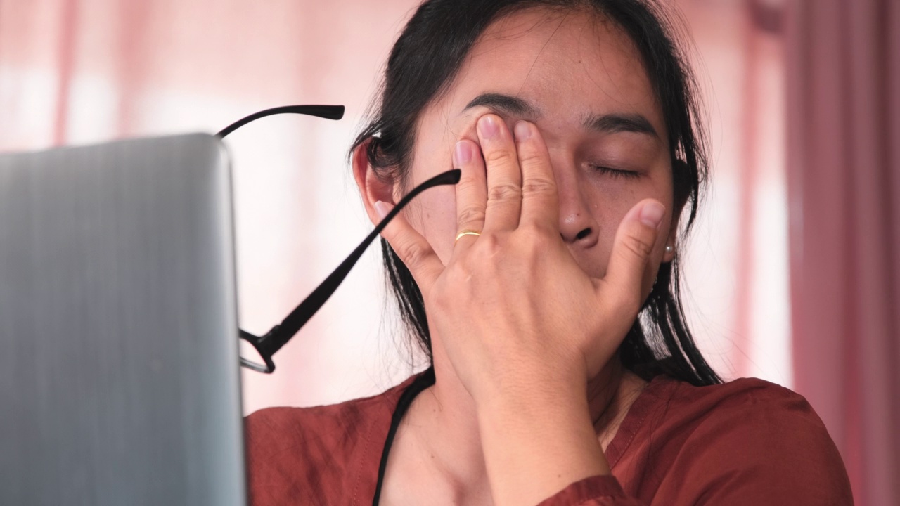 严重的亚洲女性在家里长时间使用笔记本电脑工作后感到疲劳和眼睛疲劳。健康和办公室综合症的概念。