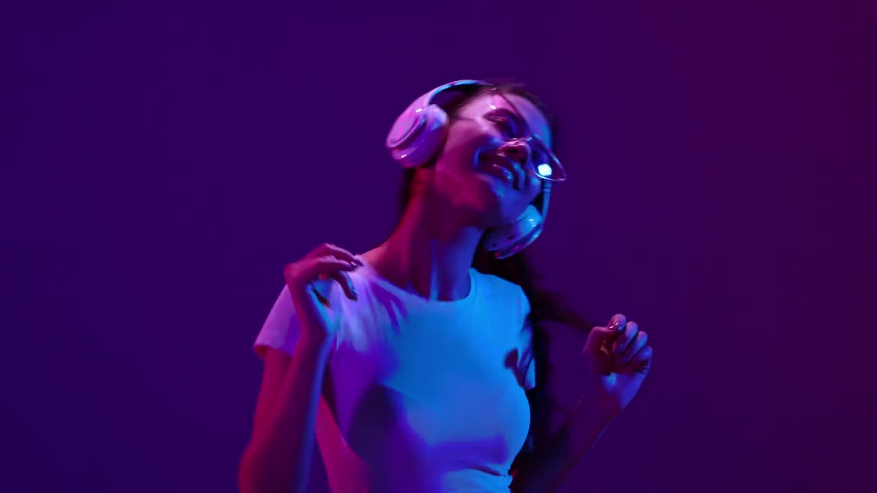 快乐微笑的年轻男女，戴着耳机听音乐，在霓虹灯滤镜的深蓝色背景下跳舞。4 k。情感、广告、文化、青春观念