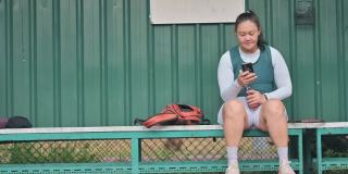 一名亚裔华裔中年女子坐在网球场的长椅上，在比赛结束后用智能手机查看自己的心跳