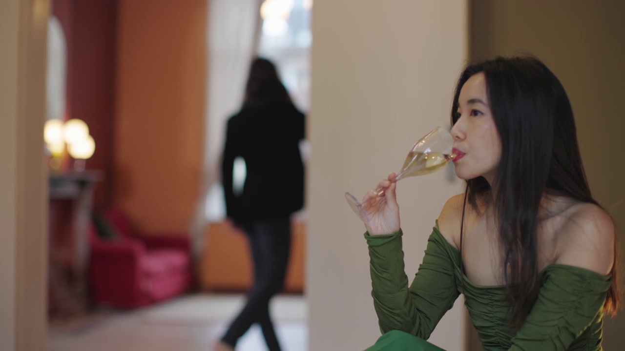 亚洲妇女在优雅的绿色连衣裙坐在床上啜饮一长笛白葡萄酒穿夹克的男人在讲电话的背景