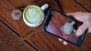 美食博主用手机拍摄热抹茶拿铁，在社交媒体、在线、数字生活方式中获取新内容。垂直拍摄。在木桌上放着一大杯热饮，上面有红心形状的泡沫