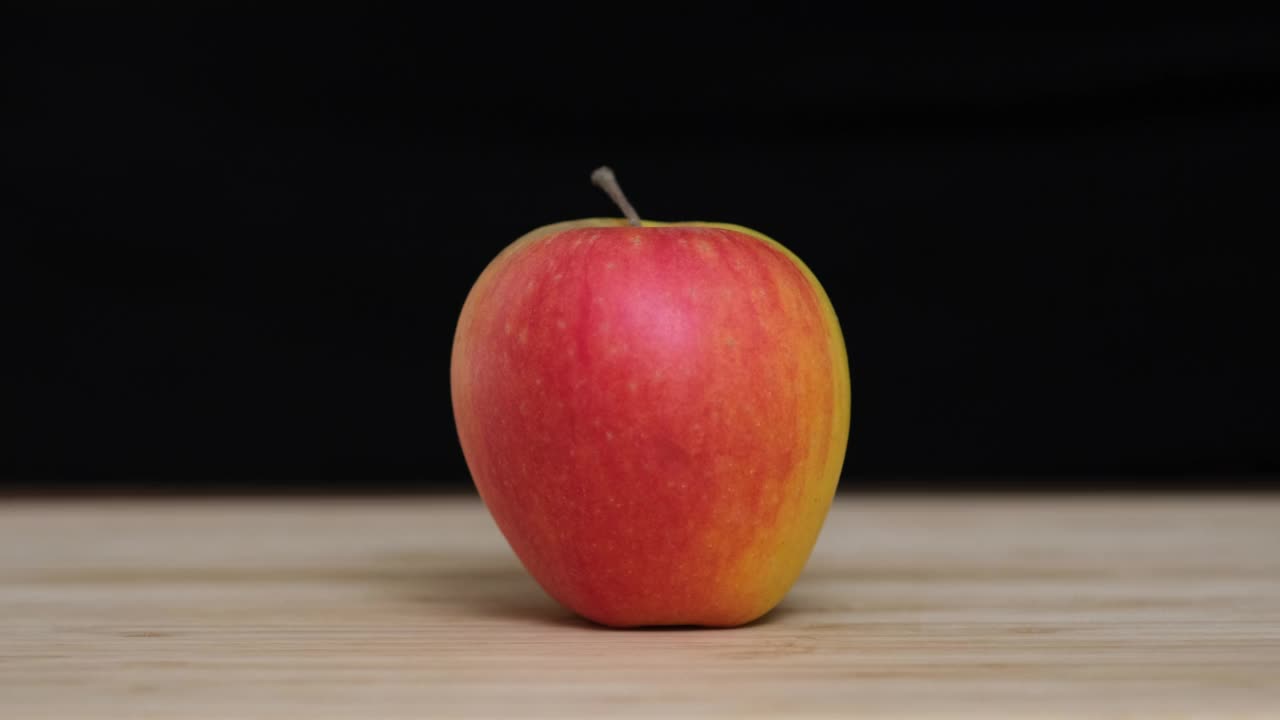 一个红苹果躺在切菜板上。一把菜刀把一个红苹果切成两半。在黑色的背景上用刀把苹果切成两半。人手戴着黑色手套切水果，特写慢动作