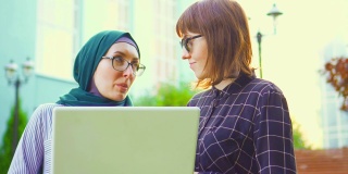 穆斯林女学生戴着头巾和眼镜，她的朋友戴着眼镜在公园里用笔记本电脑聊天，太阳耀斑。缓慢的莫