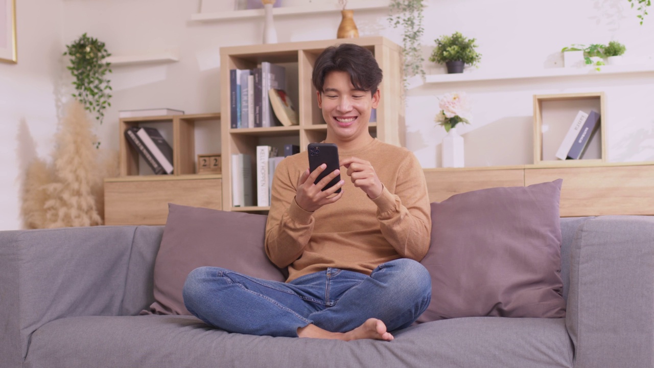 一名亚洲男子坐在家里的沙发上，用他的智能手机工作。人们在使用社交媒体应用程序时工作。在家工作的概念。