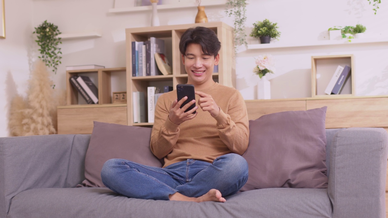 详情图-一名亚洲男子坐在家里的沙发上，用他的智能手机工作。人们在使用社交媒体应用程序时工作。在家工作的概念。