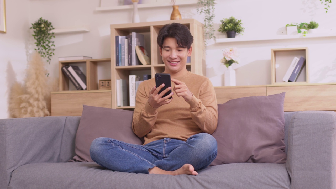 详情图-一名亚洲男子坐在家里的沙发上，用他的智能手机工作。人们在使用社交媒体应用程序时工作。在家工作的概念。