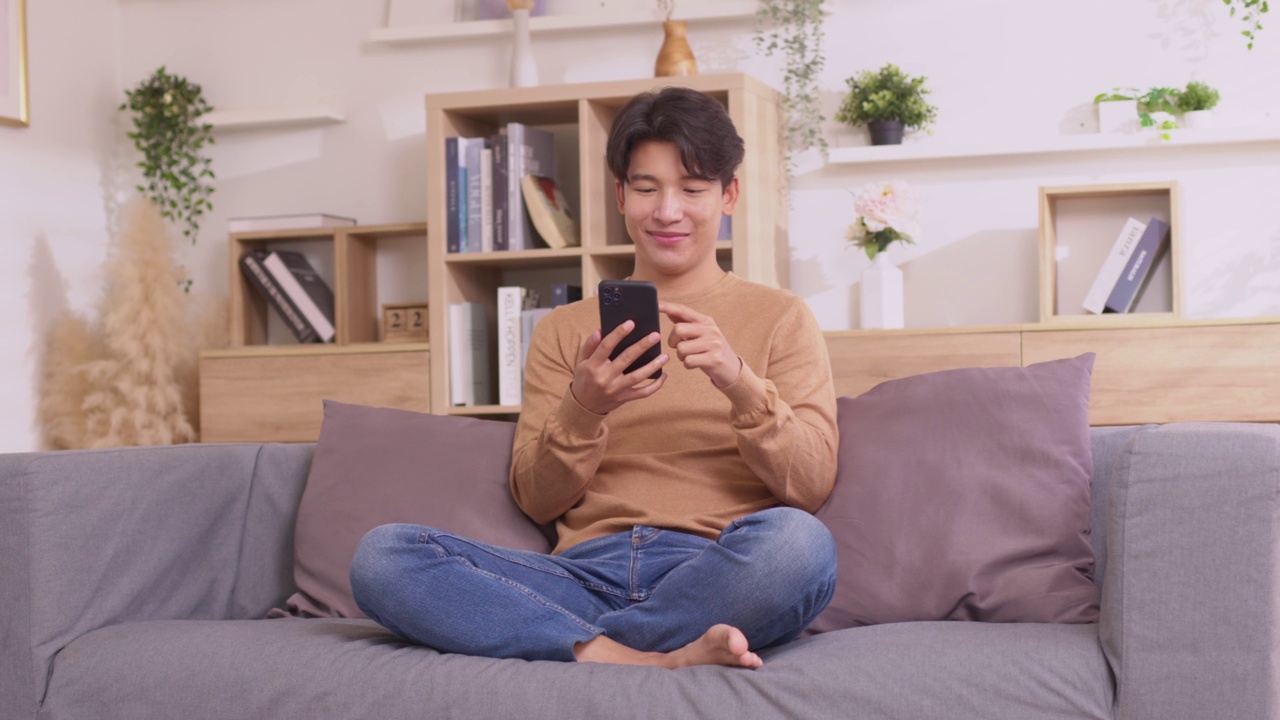 详情图-一名亚洲男子坐在家里的沙发上，用他的智能手机工作。人们在使用社交媒体应用程序时工作。在家工作的概念。