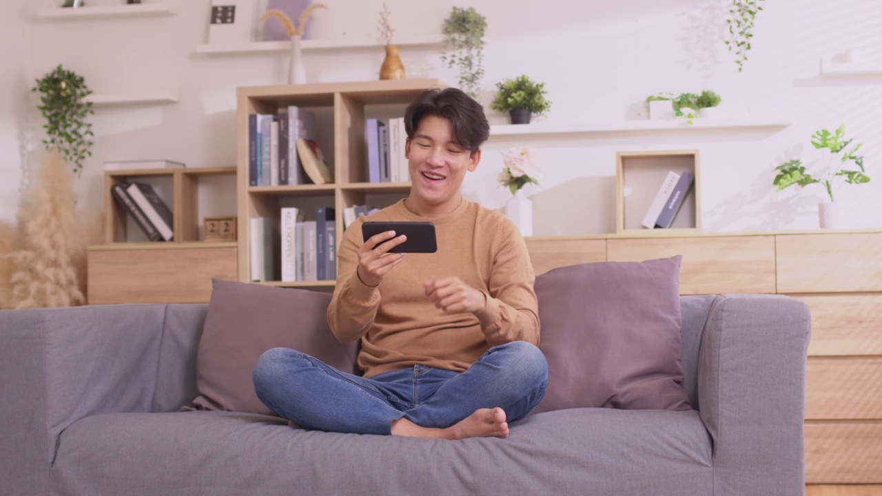 在家里的客厅里。亚洲男人早上在沙发上玩手机游戏，玩得很开心。