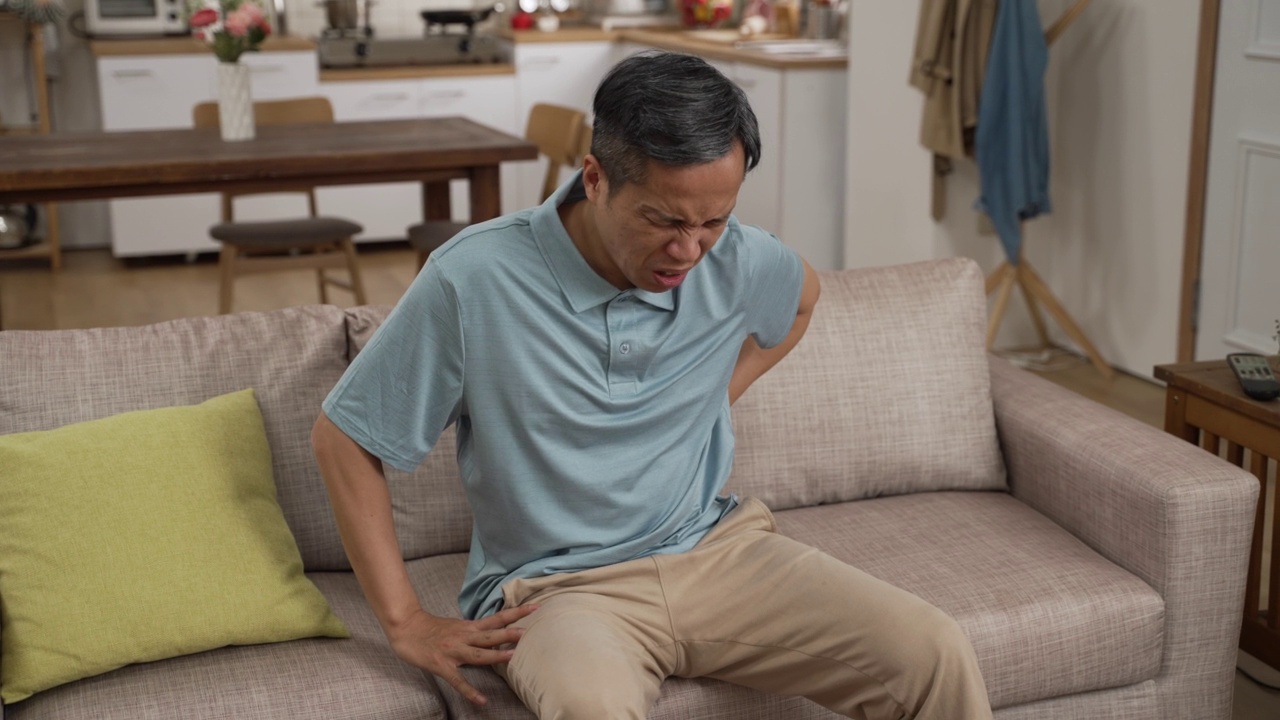 中国成熟男人坐在沙发上休息时，腰疼，呻吟，非常不舒服。老年人在家跌倒风险观念