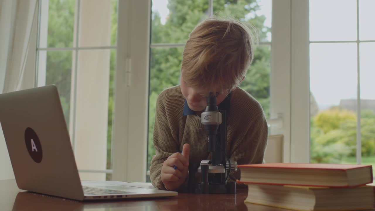 小男孩看着显微镜，对着笔记本电脑聊天，在家在线学习。家庭教育的概念。