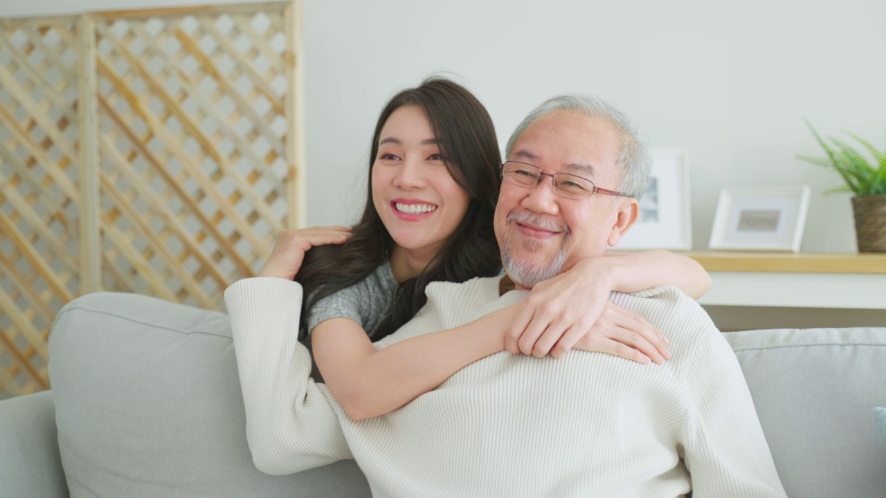 可爱的亚洲家庭，年轻的女儿问候并拥抱年长的父亲。迷人的女人孙女拜访老人成熟的男人在家里享受在客厅一起度过休闲时间和微笑。