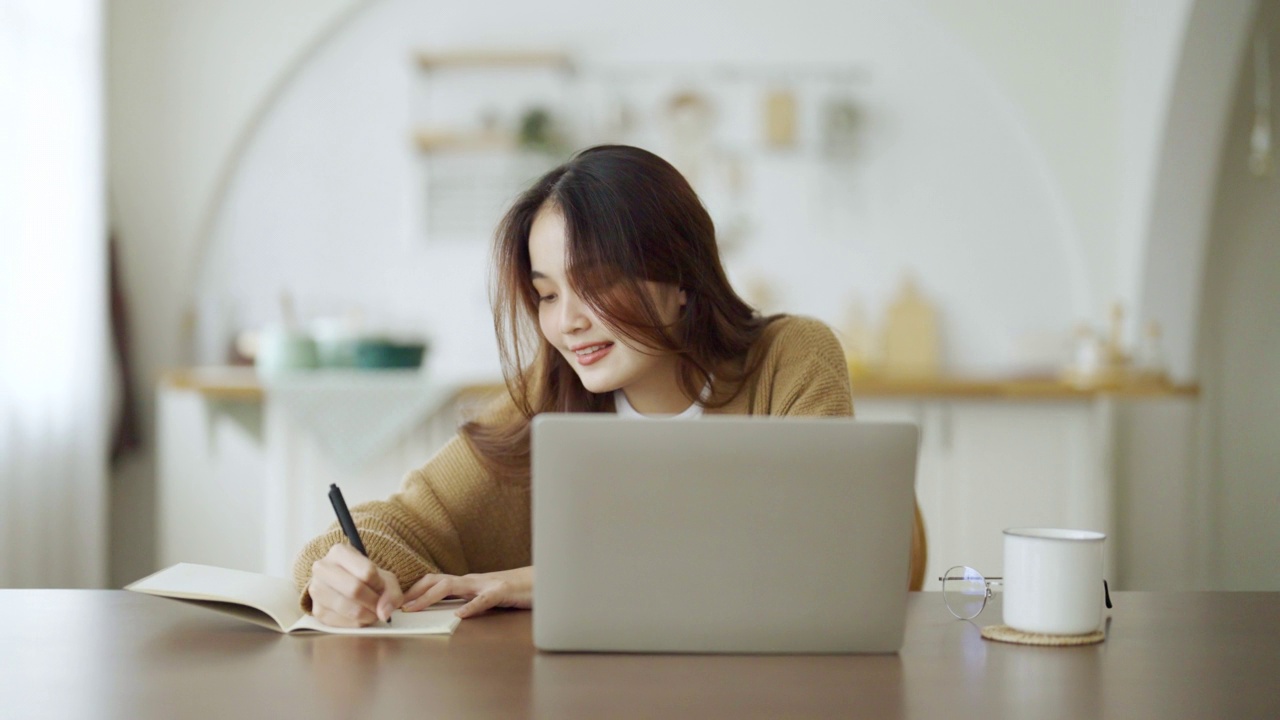 年轻的亚洲女人坐在桌子上，在笔记本上做笔记。在桌子上用笔记本电脑、文件和咖啡工作。学生学习在网上