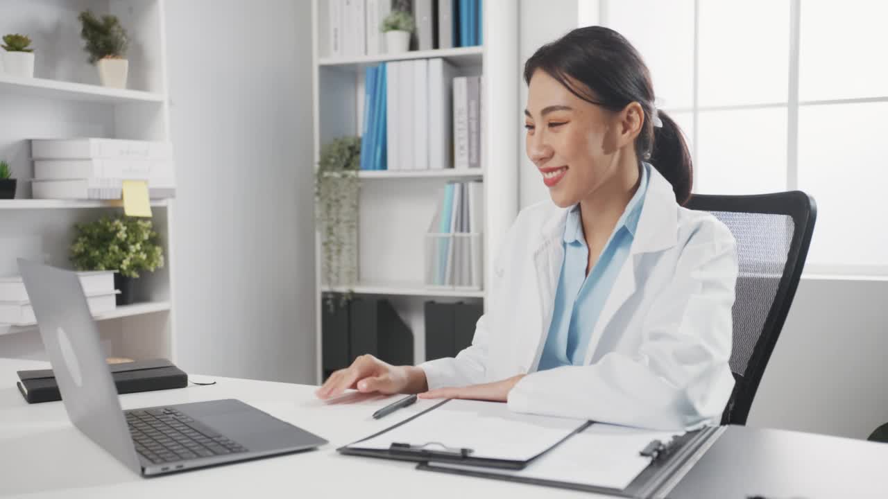 年轻的亚洲女医生穿着白色医疗制服，带着听诊器，使用电脑笔记本电脑在健康诊所或医院的办公桌上与病人进行视频电话会议。咨询和治疗理念。