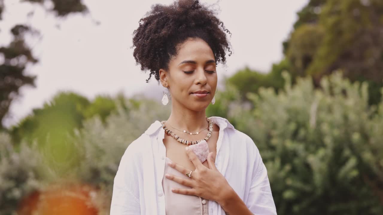 年轻的女人与卷曲的头发呼吸新鲜的空气，而冥想与一个玫瑰石英宝石独自在户外。一位女性在大自然中康复时看起来很平静，感觉很满足