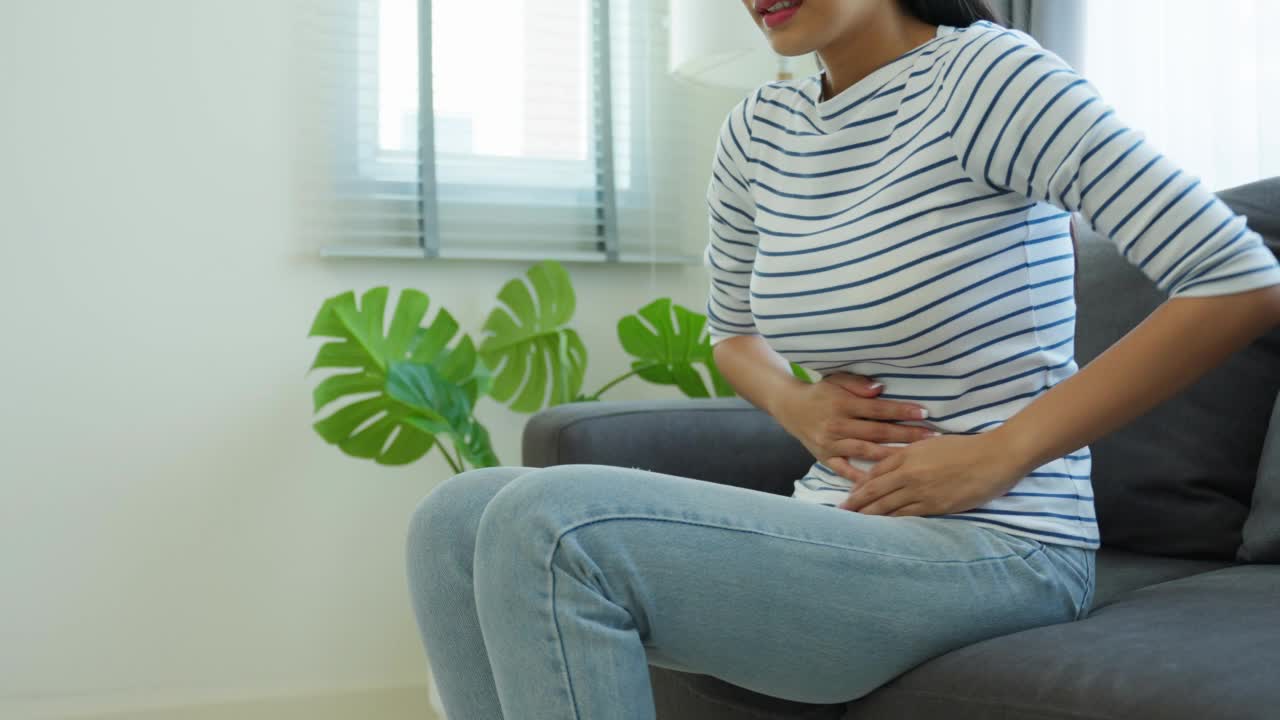 亚洲美丽的生病的女孩遭受伤害胃痛在客厅。有魅力的年轻女性在床上醒来后会感到难受和疼痛，胃痛或月经来了，然后在早上抱着肚子