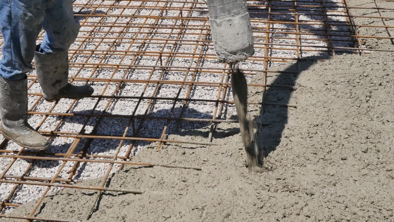 一名穿着大型防水靴的建筑工人将混凝土砂浆灌入地板上的钢筋中，并将一根宽软管连接到供应泥浆的混凝土泵的套筒上。基础混凝土的