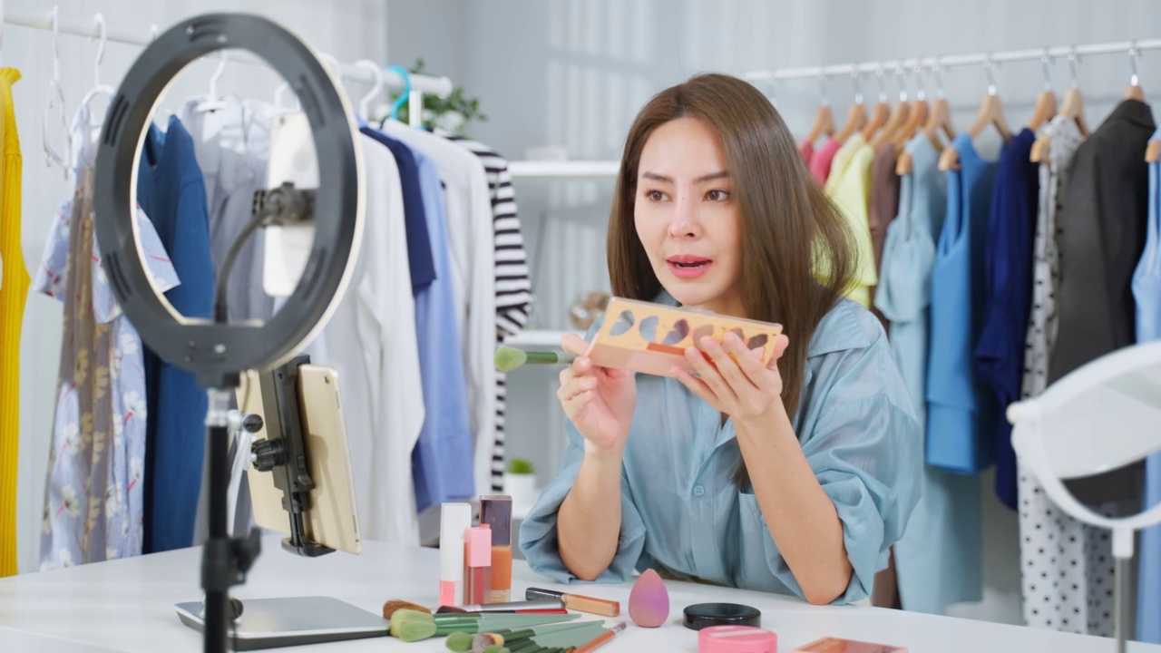 詳情圖-亞洲女性視頻博主與相機(jī)交談，回顧美容在家里。美妝博主網(wǎng)紅女孩坐在沙發(fā)上直播，向室內(nèi)的粉絲展示眼影調(diào)色板。