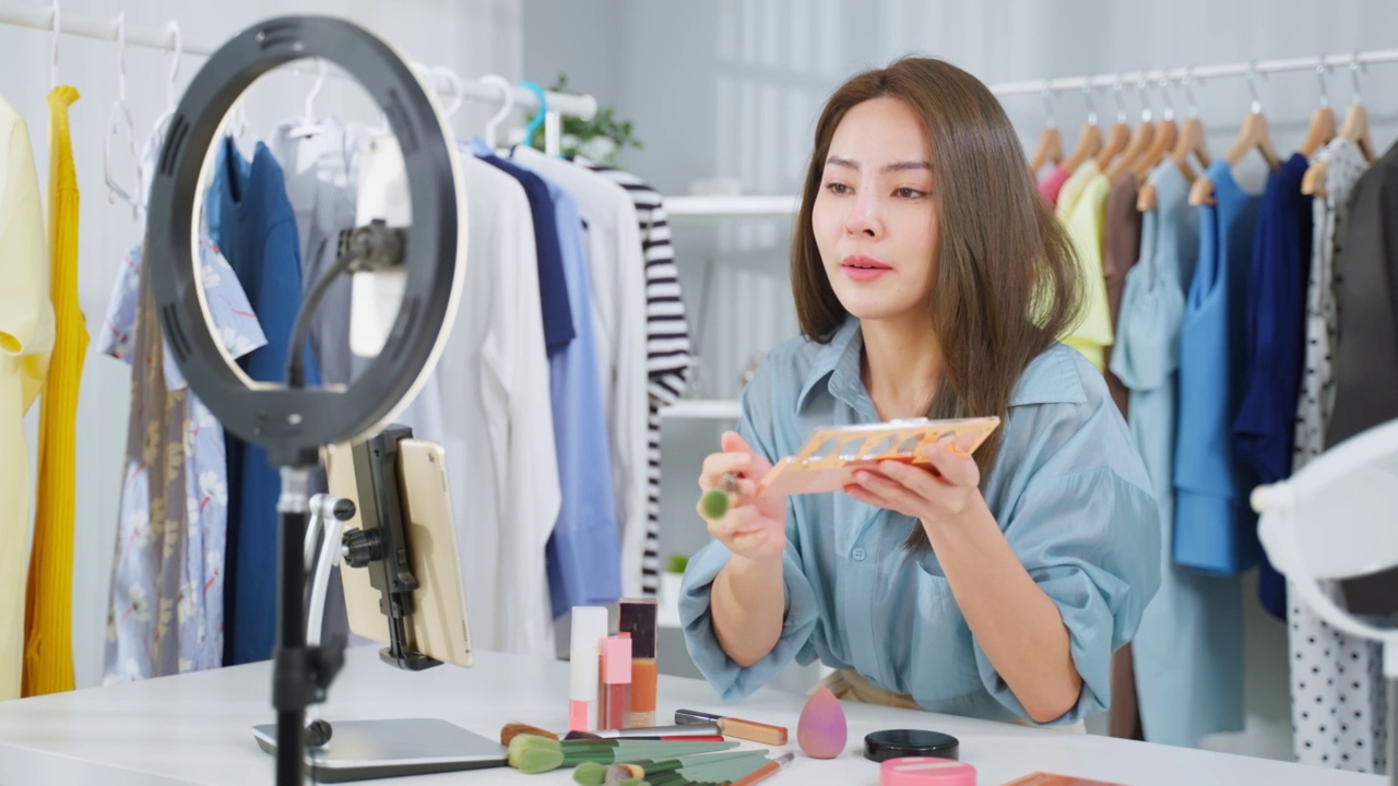詳情圖-亞洲女性視頻博主與相機(jī)交談，回顧美容在家里。美妝博主網(wǎng)紅女孩坐在沙發(fā)上直播，向室內(nèi)的粉絲展示眼影調(diào)色板。