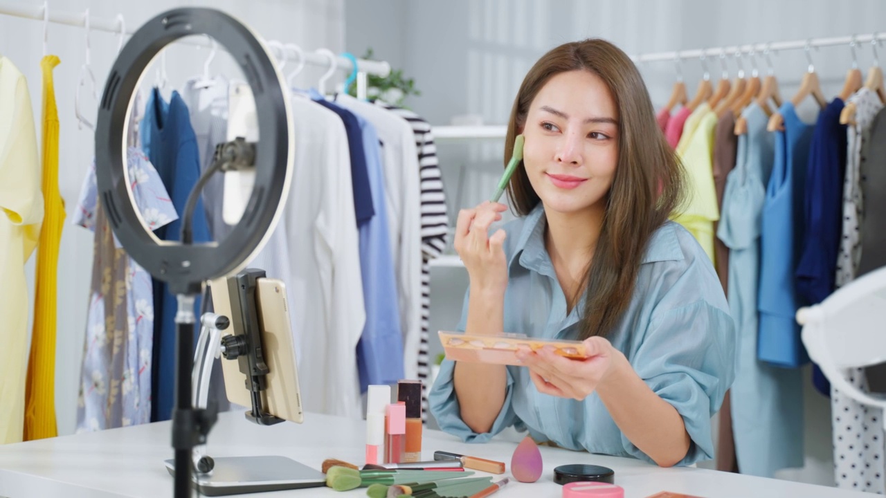 詳情圖-亞洲女性視頻博主與相機(jī)交談，回顧美容在家里。美妝博主網(wǎng)紅女孩坐在沙發(fā)上直播，向室內(nèi)的粉絲展示眼影調(diào)色板。