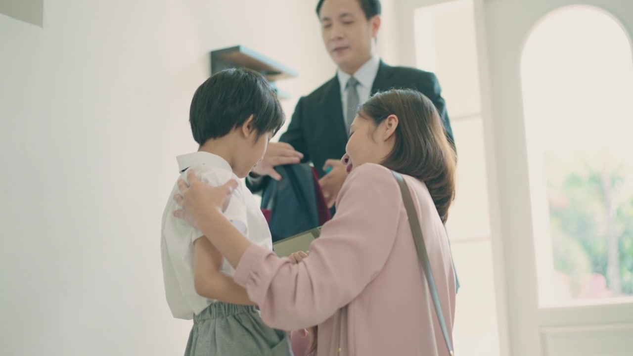 詳情圖-亞洲媽媽抱著她的小兒子，準(zhǔn)備送他去上學(xué)，而亞洲爸爸在家里的客廳里背著兒子的書(shū)包，準(zhǔn)備去上學(xué)。親子關(guān)系或愛(ài)與紐帶表達(dá)的概念。